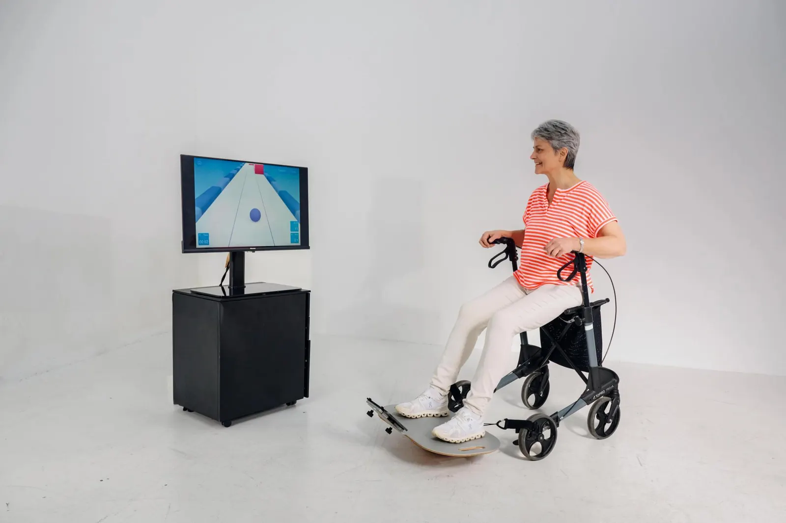 Seniorin sitzt auf ihrem Rollator und steuert mit den Füßen auf dem I.S.T. Board ein Spiel am Bildschirm